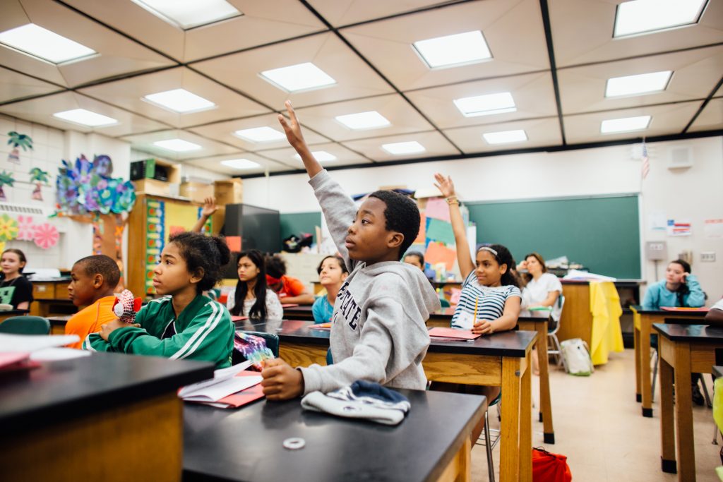 Young boy at front desk raises hand high in public school room full of students